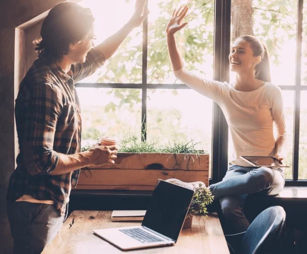 Nasmejan moški in ženska si dajeta petko ob oknu v svetli pisarni ali kavarni kjer na mizi ležita prenosnik in beležka kar kaže na uspešen trenutek sodelovanja ali dosežen cilj
