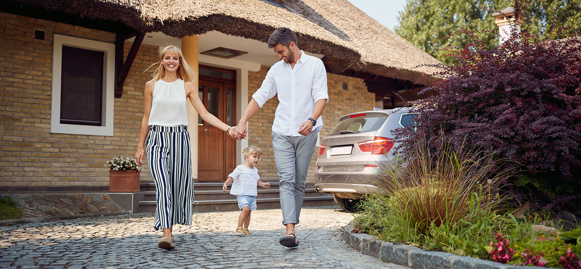 Nasmejana družina z majhnim otrokom hodi po dovozu pred hišo z avtom in slamnato streho