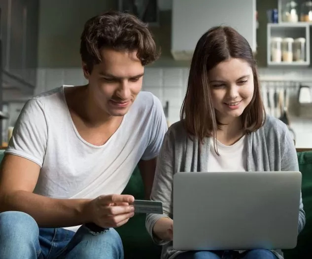 Mlajši par sedi na kavču, dekle ima pred seboj prenosni računalnik, fant pa drži v roki kreditno kartico