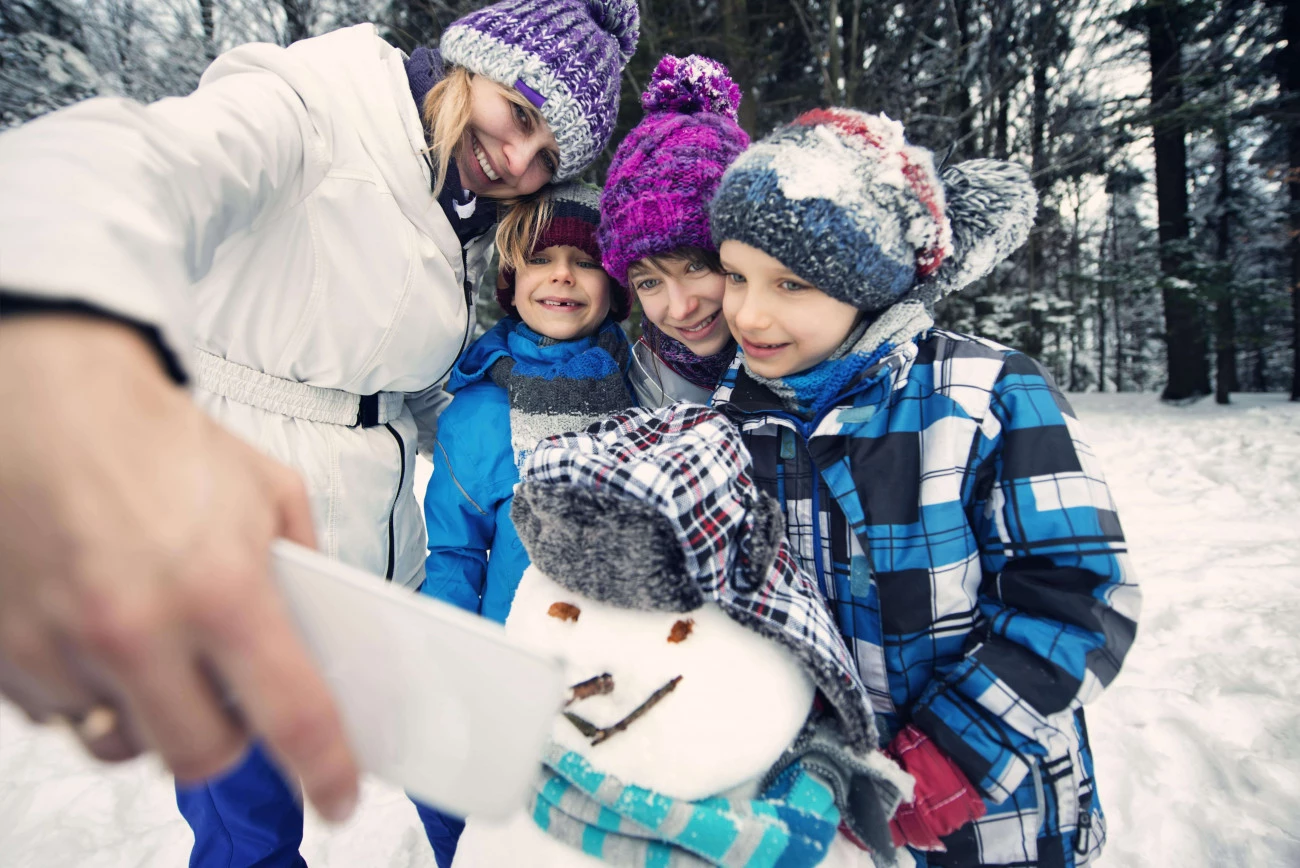 Družina oblečena v zimska oblačila z nasmehi dela selfi ob snežaku v zasneženem gozdu
