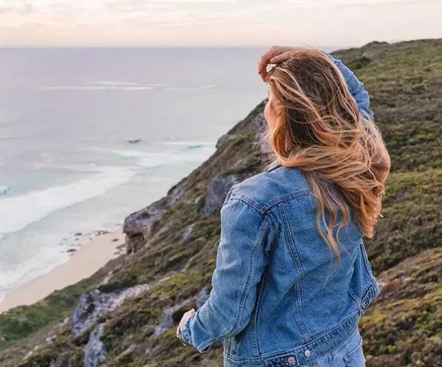Ženska v jeans jakni stoji na klifu in gleda proti oceanu z vetrom v laseh