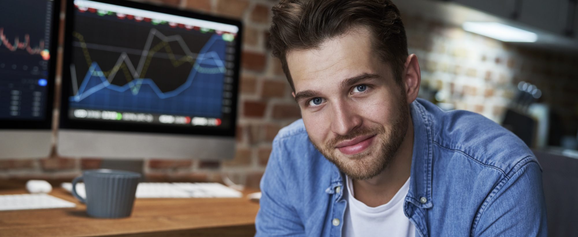 Nasmejan moški v modri srajci sedi pred dvema zaslonoma z grafi finančnih trgov v domači pisarni