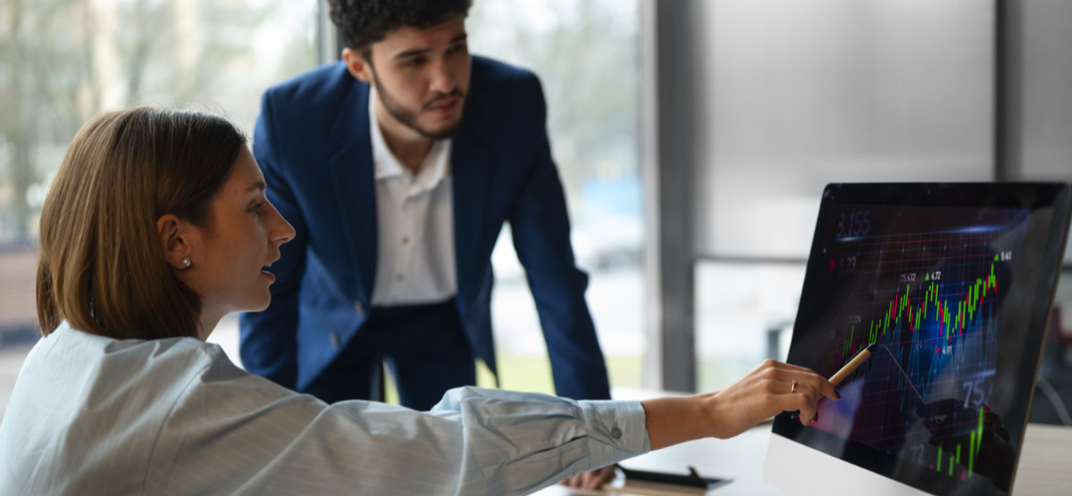 Ženska sedi za računalnikom in s svinčnikom kaže na rast finančnega grafa medtem ko moški v obleki stoji ob njej in opazuje