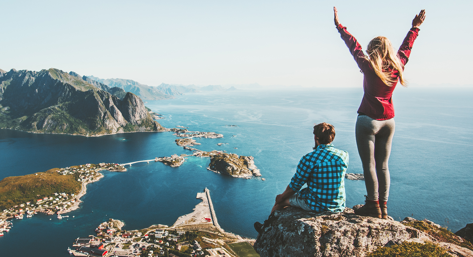 Par stoji na vrhu klifa z razgledom na fjord otoke in morje medtem ko ženska dviguje roke v zmagoslavju