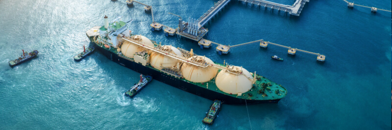 Zračni pogled na industrijsko pristanišče ob morju, kjer velik tanker za utekočinjen plin (LNG) pristaja ob pomolu, obdan z več manjšimi vlečnimi ladjami; v ozadju so rezervoarji, industrijski objekti in obalna infrastruktura.