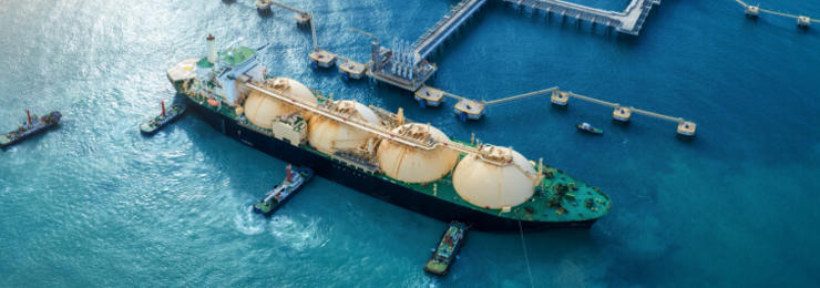 Zračni pogled na industrijsko pristanišče ob morju, kjer velik tanker za utekočinjen plin (LNG) pristaja ob pomolu, obdan z več manjšimi vlečnimi ladjami; v ozadju so rezervoarji, industrijski objekti in obalna infrastruktura.