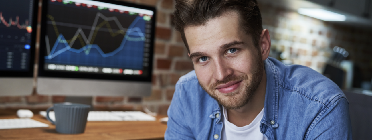 Slika prikazuje finančnega strokovnjaka ob pisalni mizi, na kateri sta računalniška monitorja s prikazom gibanja vrednosti finančnih instrumentov