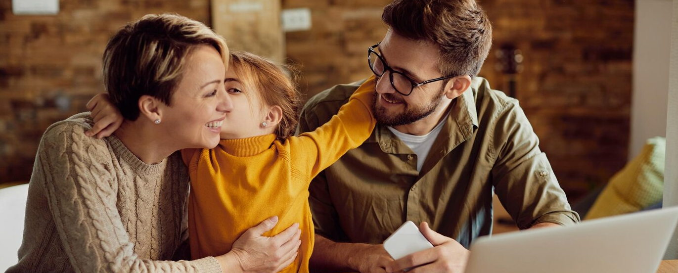 Nasmejana družina z majhno hčerko sedi doma in se objema ob uporabi prenosnega računalnika in pametnega telefona