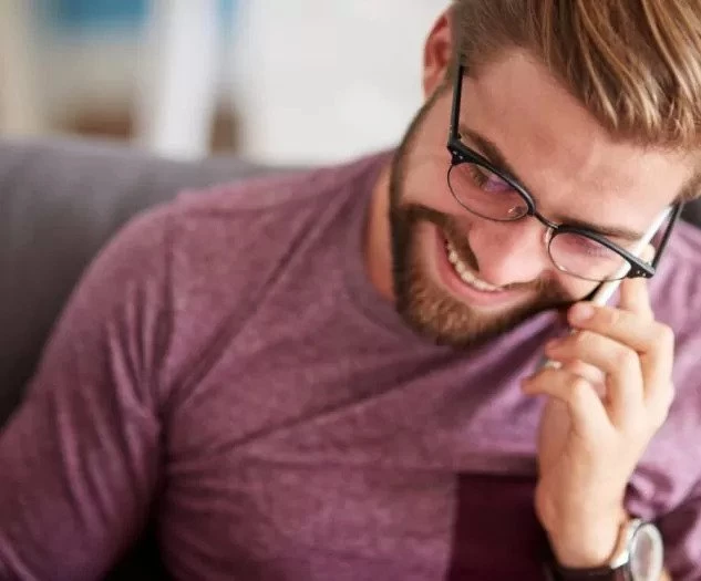 Nasmejan moški z očali in brado nosi vijolično majico in se sproščeno pogovarja po telefonu medtem ko sedi na kavču