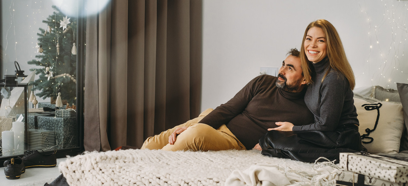 Srečen par se objema in nasmejano sedi na postelji v praznično okrašeni sobi z božičnim drevescem in darili v ozadju