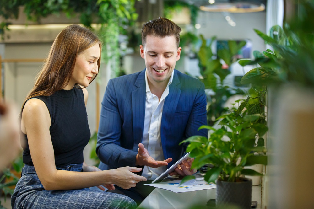 Moški in ženska sedita med zelenimi rastlinami in se nasmejana pogovarjata ob pogledu na tablico