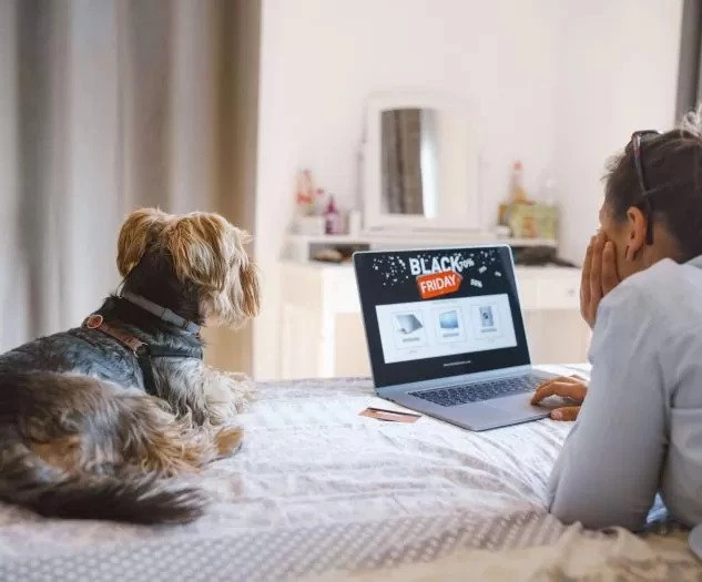 Ženska s psom leži na postelji in gleda v prenosnik z oglasom za Black Friday medtem ko ob njej leži plačilna kartica