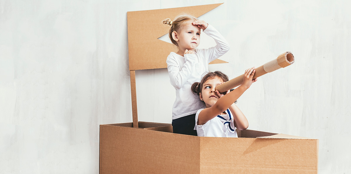 Dva otroka se igrata v kartonskem čolnu eden gleda skozi daljnogled drugi se razgleduje naprej kot da sta na pustolovščini