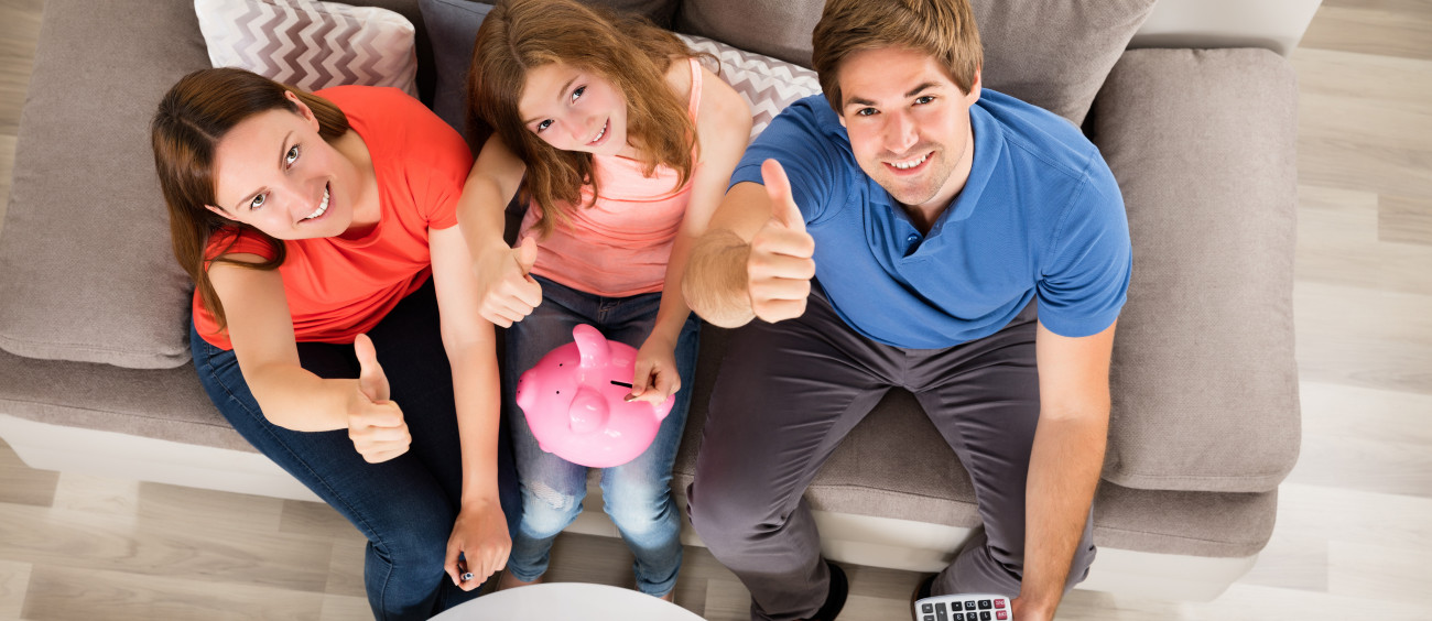 Nasmejana družina s hčerko ki v hranilnik v obliki pujska vstavlja kovanec dviguje palce v znak zadovoljstva s finančnim varčevanjem