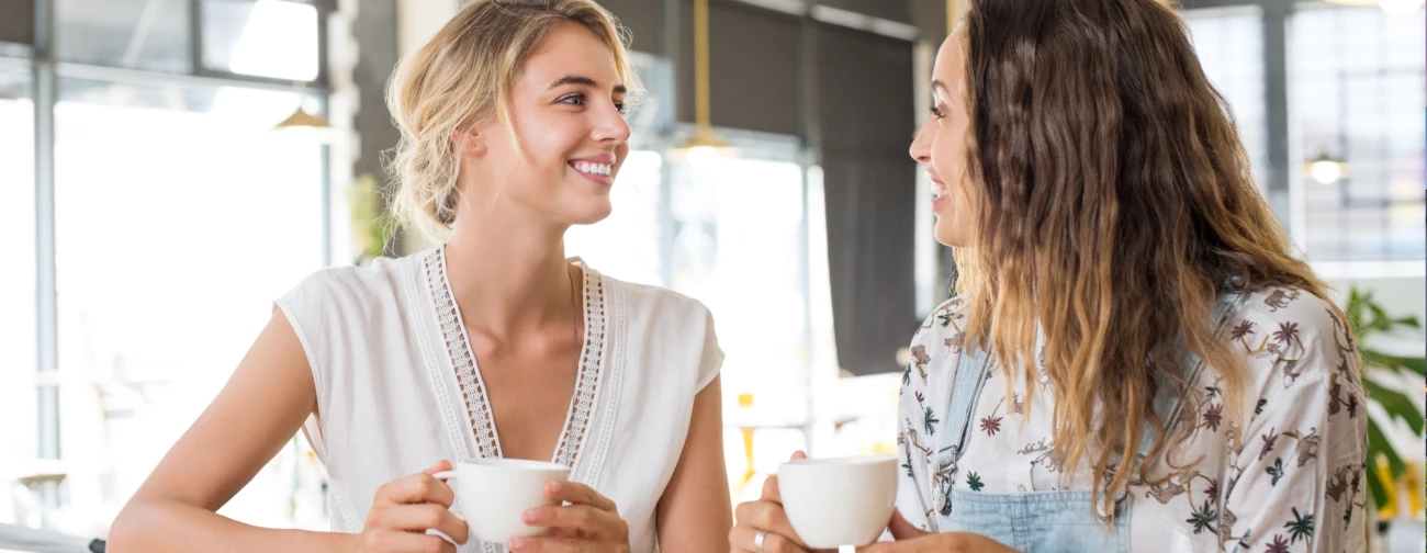 Dve Mladi Zenski V Svetlem Prostoru Ob Kavi Sprosceno Klepetata In Se Smehljata
