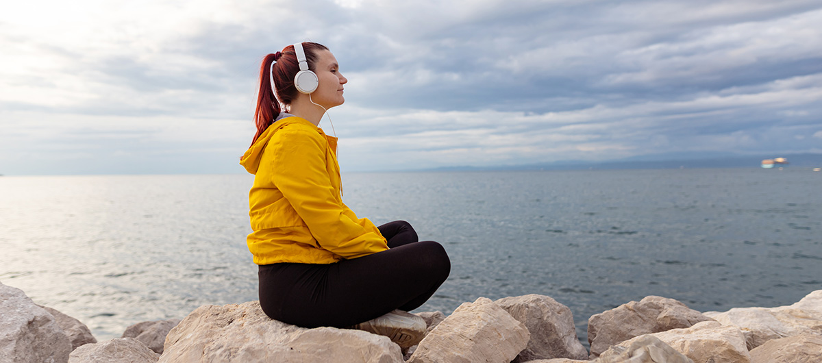 Ženska v rumeni jakni sedi na skalah ob morju in z zaprtimi očmi posluša glasbo skozi slušalke ter uživa v miru narave