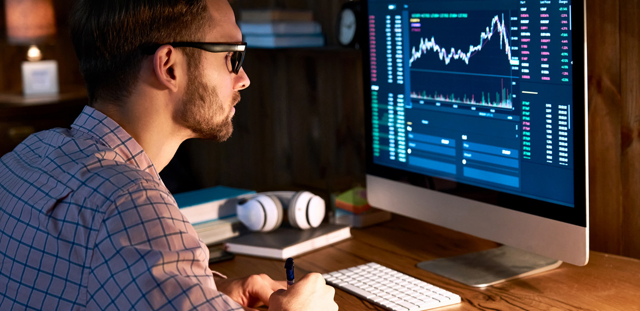 Moški z očali sedi za računalnikom in spremlja borzne grafe na zaslonu v domači pisarni
