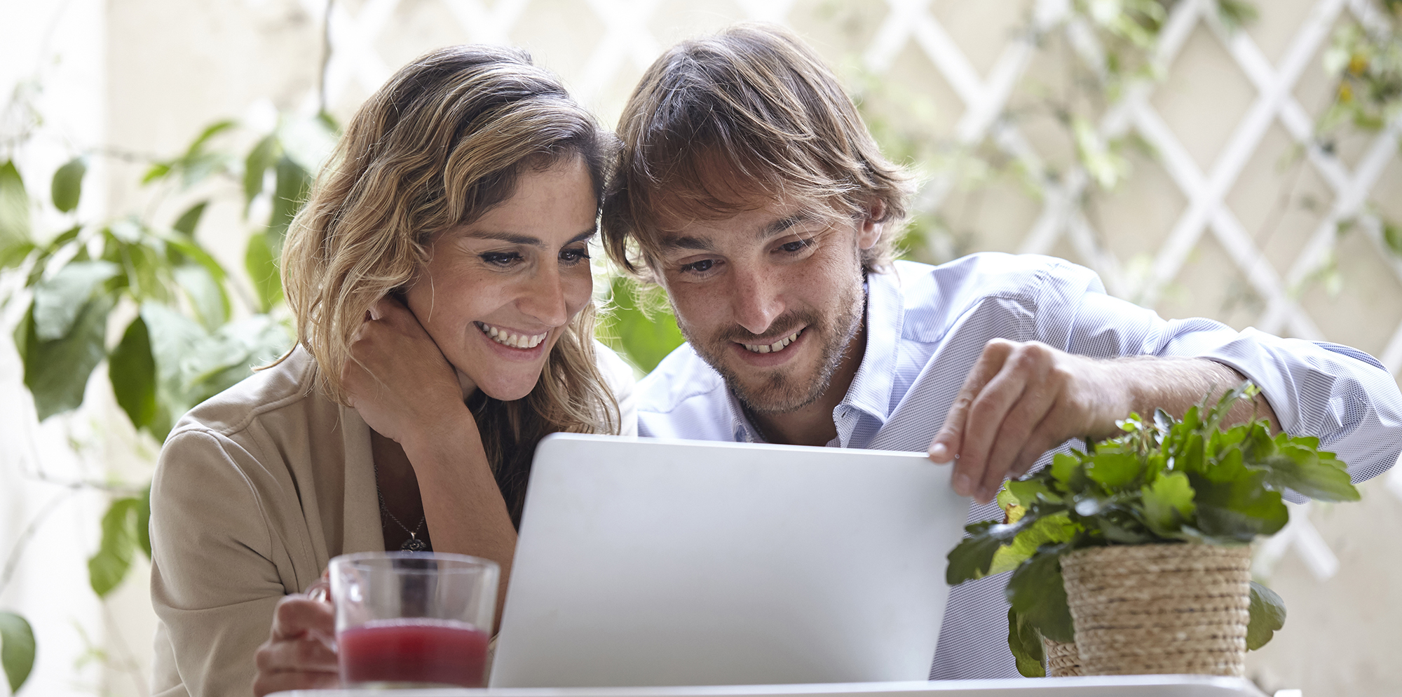 Nasmejan par skupaj gleda v prenosnik na zunanji mizi ob pijači in lončnici v prijetnem zelenem okolju