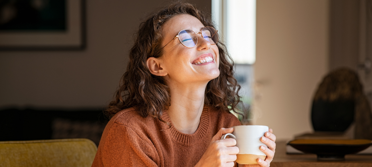 Nasmejana ženska z očali in skodelico v rokah uživa v toplem napitku medtem ko gleda navzgor z zaprtimi očmi