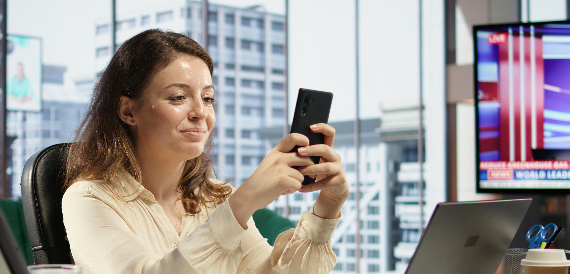 Oseba v pisarni s televizorjem, v rokah ima mobilni telefon