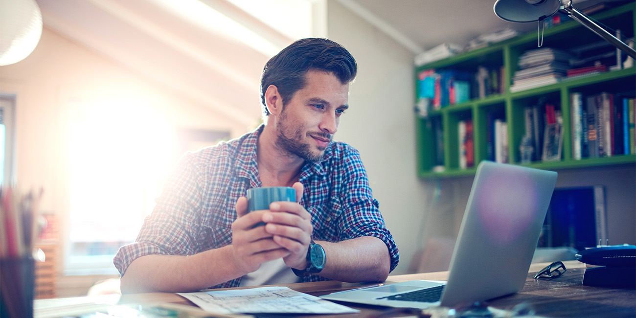 Moški sedi za mizo, v rokah ima skodelico, in gleda v zaslon prenosnega računalnika