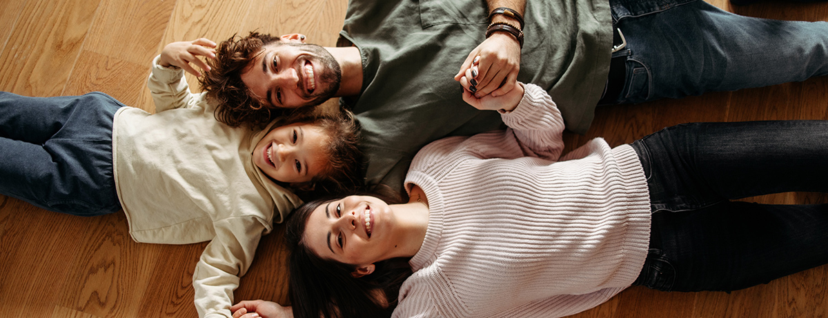 Nasmejana družina leži na lesenih tleh doma in se drži za roke ter gleda proti kameri