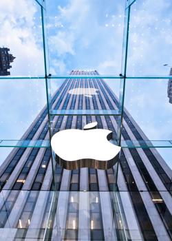 Pogled navzgor skozi stekleno streho razkriva velik bel logotip podjetja Apple in visok poslovni nebotičnik med stolpnicami v mestu