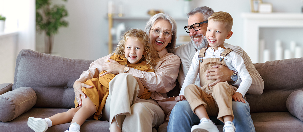 Starejši par z vnukinjo in vnukom veselo sedi na kavču v svetli dnevni sobi
