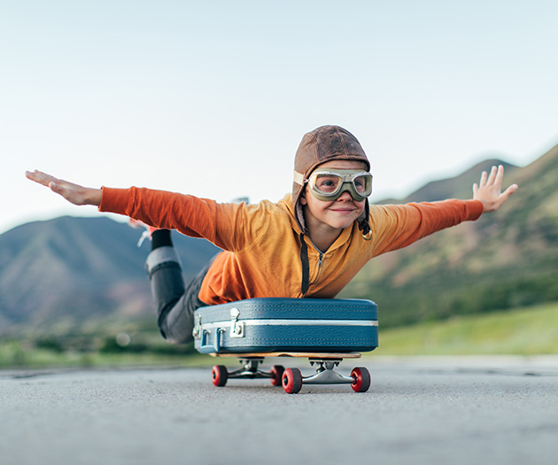 Otrok s pilotsko kapo in pilotskimi očali se z razprtimi rokami pelje na rolki, tako da leži na njej