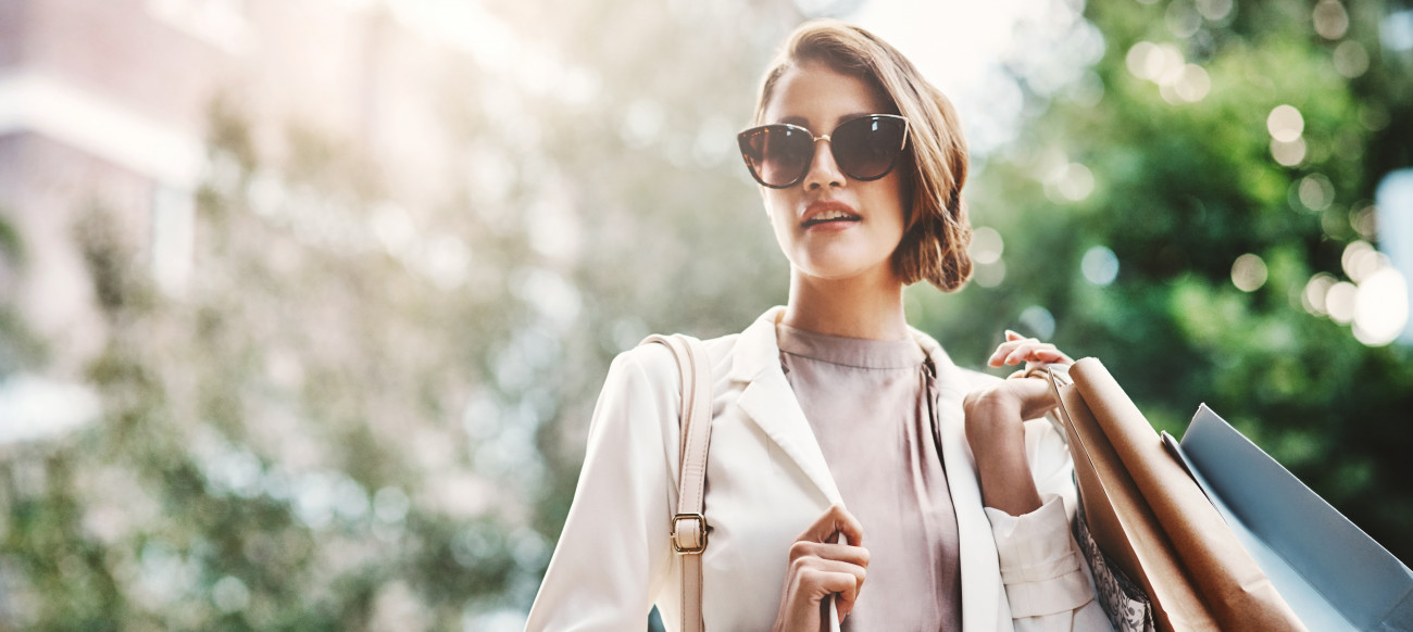 Elegantna ženska z očali za sonce in nakupovalnimi vrečkami samozavestno hodi po mestu v sončnem dnevu