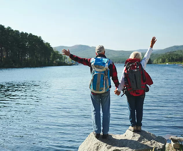 Upokojeni par stoji na skali ob jezeru in z dvignjenimi rokami zre v daljavo