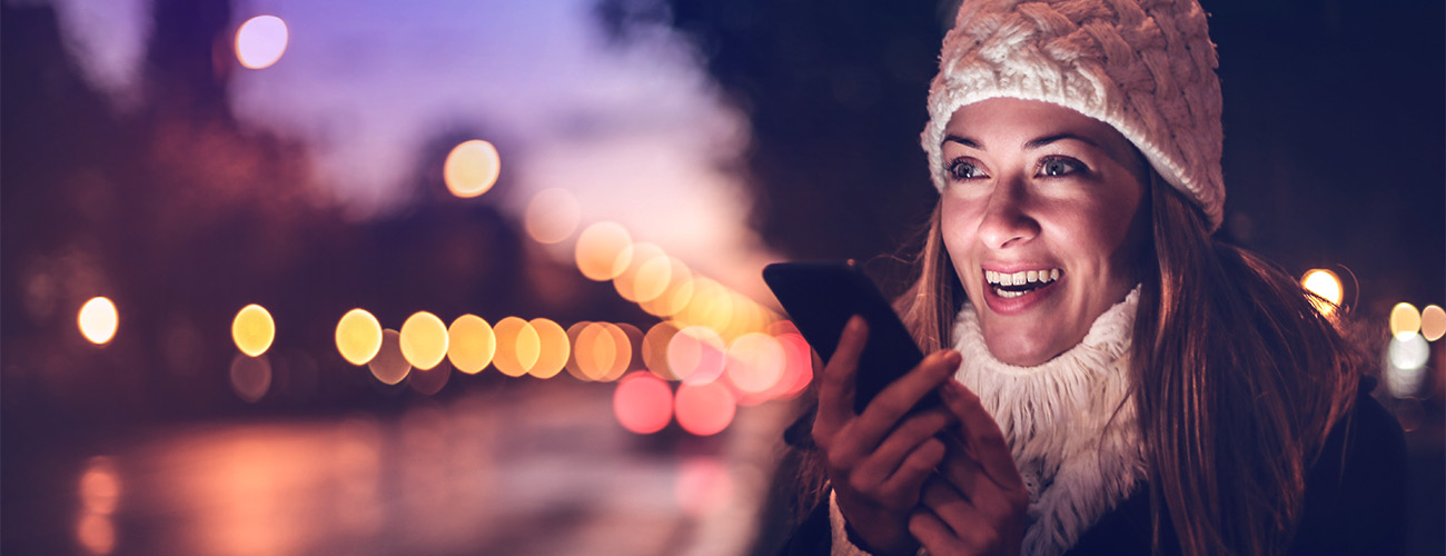 Slika ženske na ulici ob sončnem zahodu, pred obrazom drži mobilni telefon.