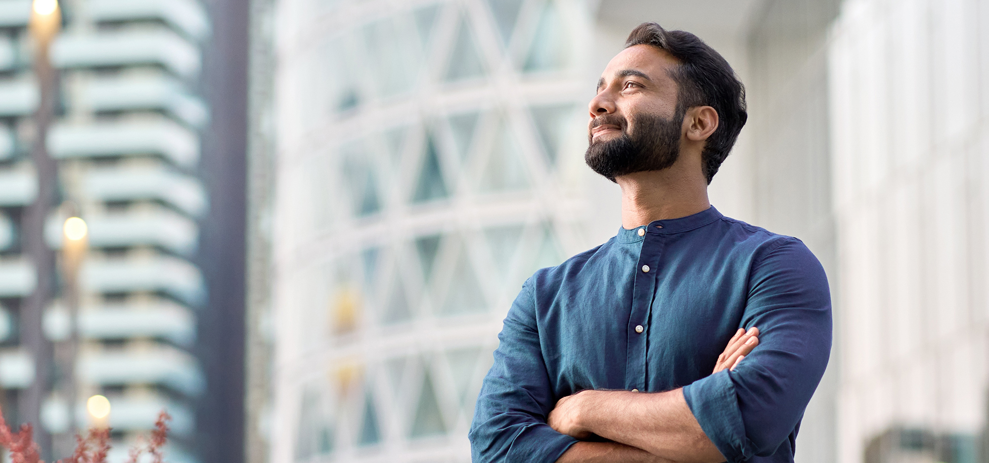 Moški z brado samozavestno stoji z zloženimi rokami in gleda navzgor pred sodobno mestno arhitekturo