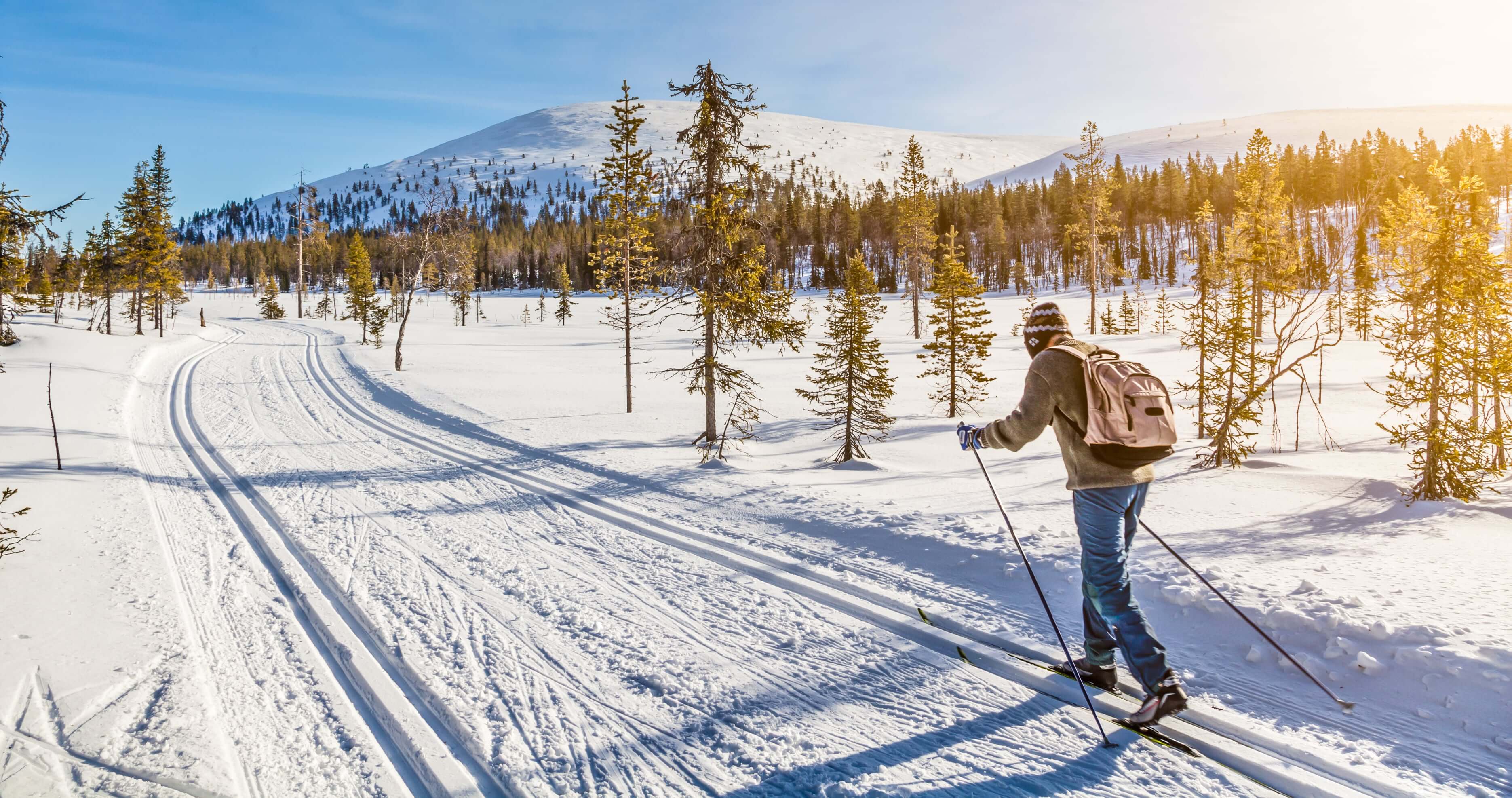 Oseba s pohodniškim nahrbtnikom teče na smučeh po urejeni snežni progi skozi zasneženo pokrajino z redkimi drevesi in gorami v ozadju