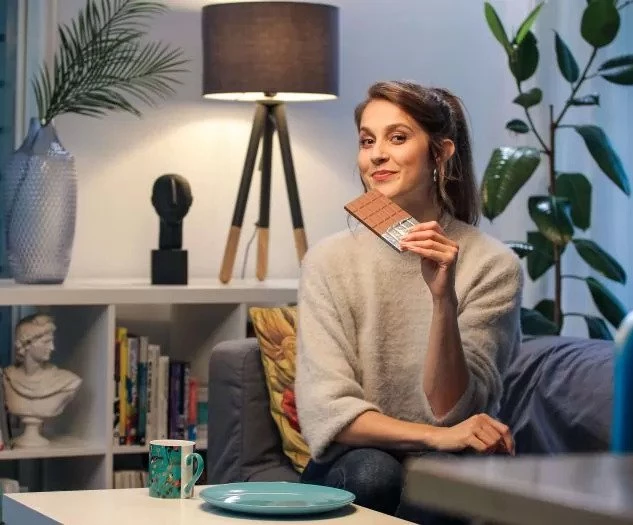 Ženska sedi sproščeno na kavču v prijetno osvetljenem dnevnem prostoru in z nasmehom drži tablico čokolade ob pripravljeni skodelici in krožniku