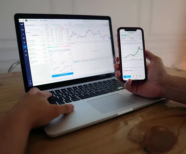 Oseba sedi za mizo in spremlja borzne grafe na prenosniku ter hkrati drži pametni telefon z enakimi podatki kar ponazarja aktivno trgovanje na več napravah