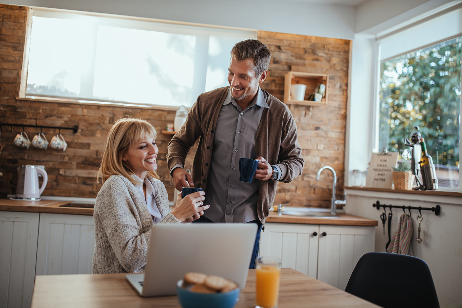 Par srednjih let v kuhinji ob kavi in prenosniku, sproščen pogovor ob domačem okolju