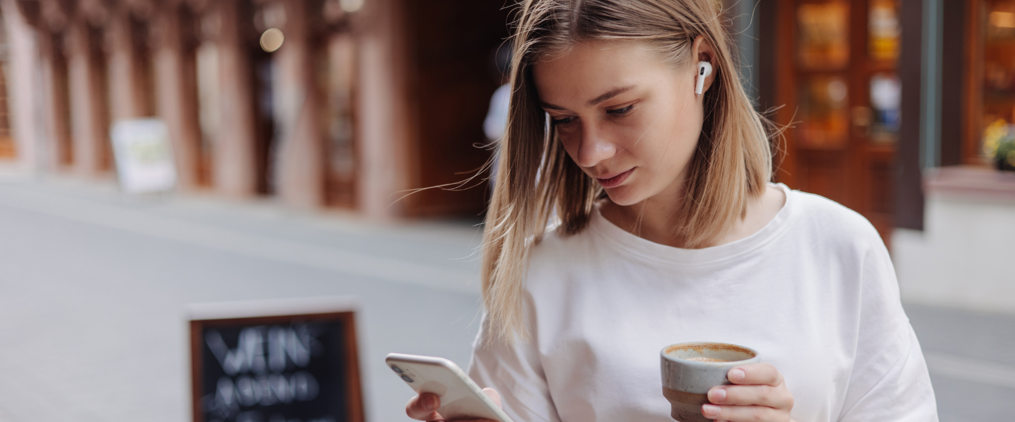 Oseba stoji na ulici in gleda v mobilni telefon, v drugi roki ima skodelico kave