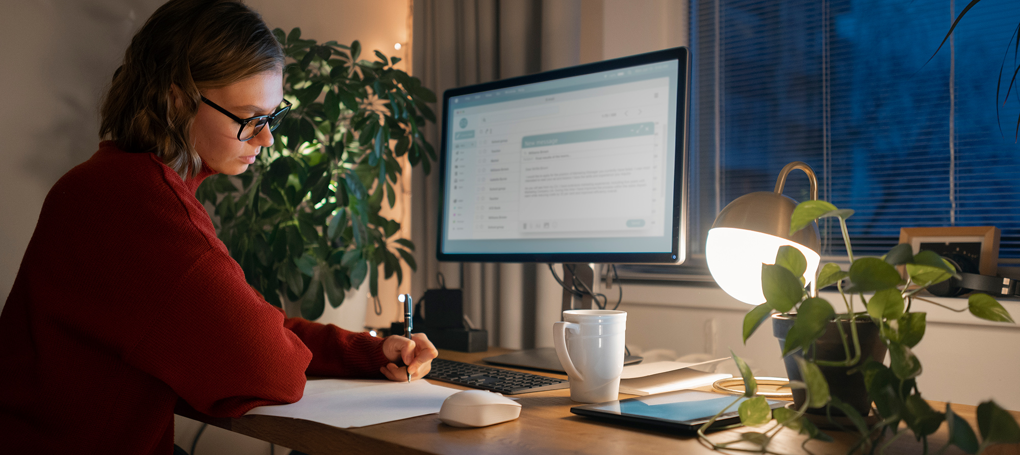 Ženska z očali dela za računalnikom in piše zapiske ob skodelici čaja v toplo osvetljeni sobi