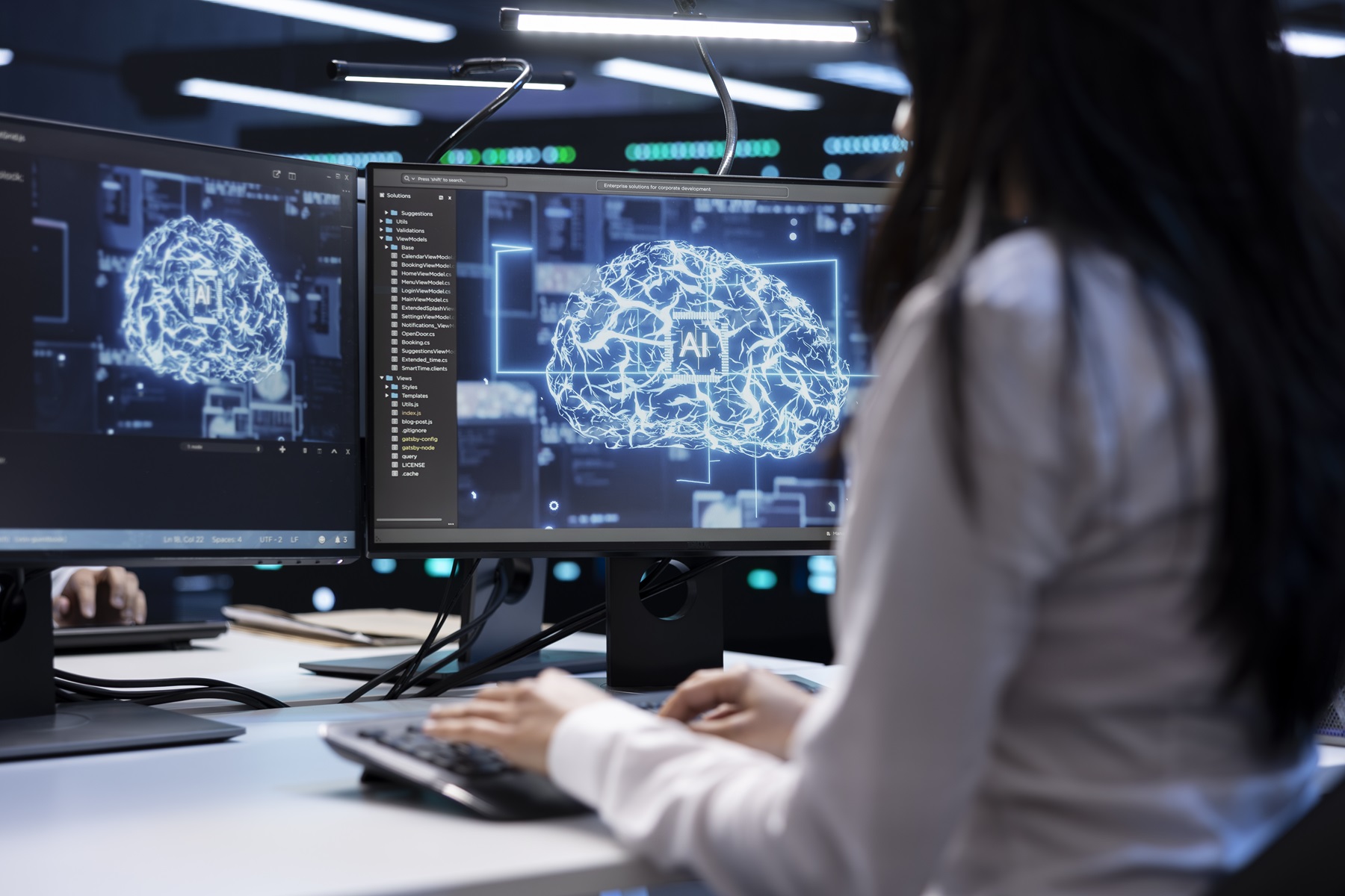 Ženska sedi pred dvema zaslonoma z digitalnim prikazom umetne inteligence v obliki osvetljenih možganov in tipka na tipkovnico 