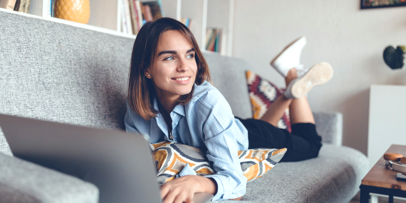 Mlajša ženska leži na kavču, pred seboj ima prenosni računalnik