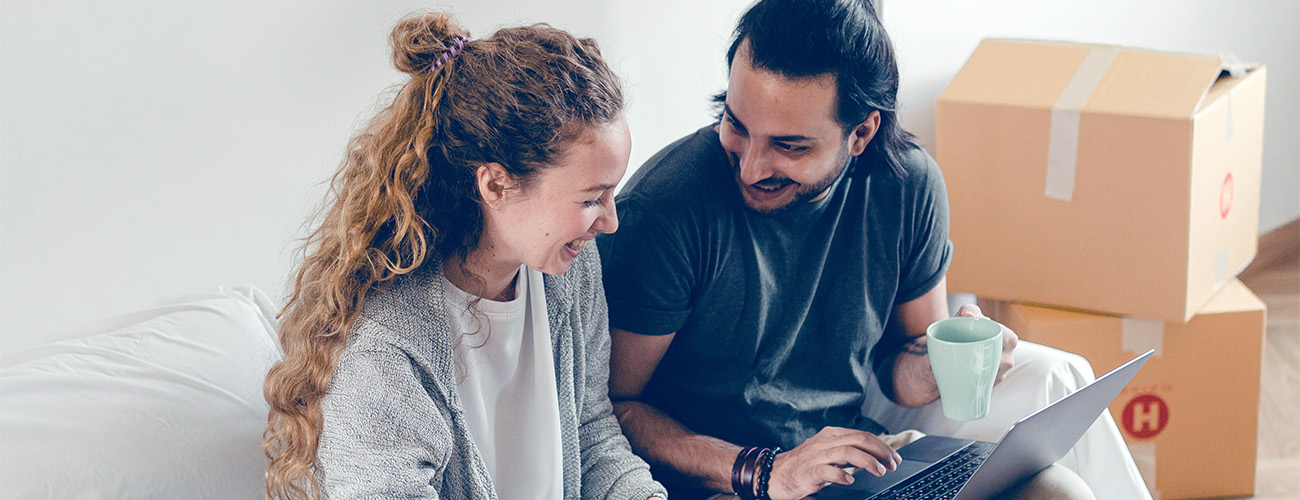 Par v tridesetih sedi na kavču v novem stanovanju, v ozadju so selitvene škatle.