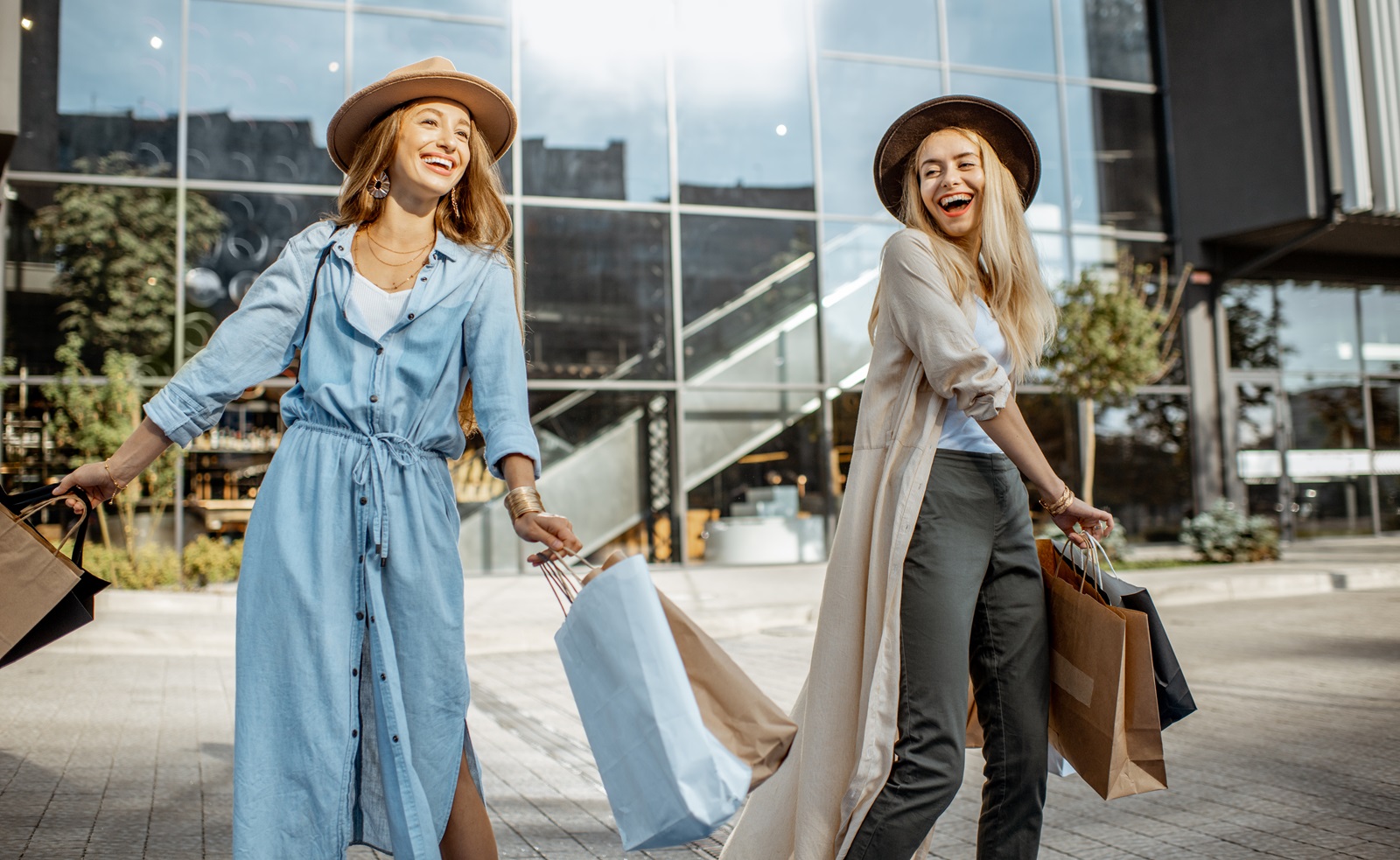 Nasmejani prijateljici v klobukih stojita pred sodobnim trgovskim centrom in držita nakupovalne vrečke v rokah