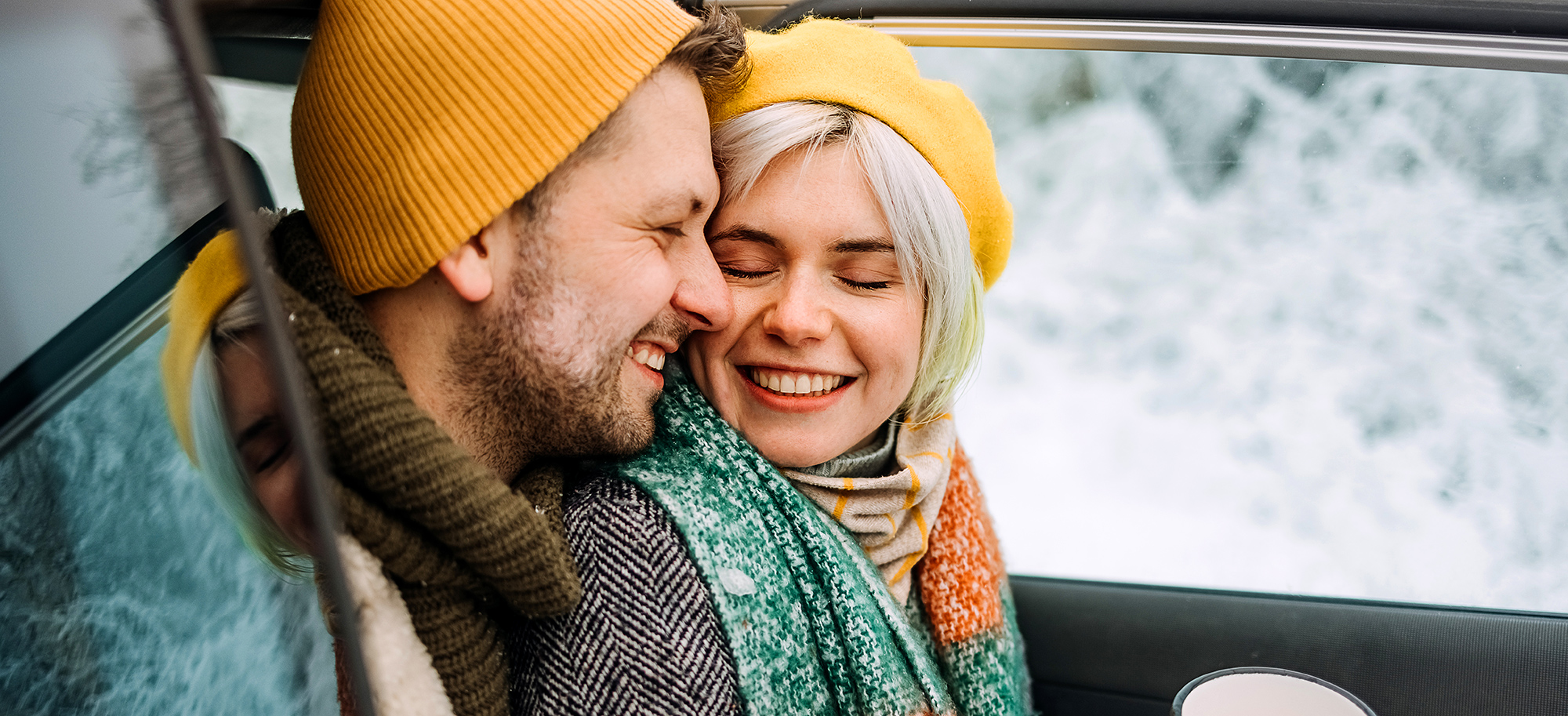 Par v zimskih oblačilih se smeji in objema ob odprtih vratih avtomobila z zasneženim ozadjem