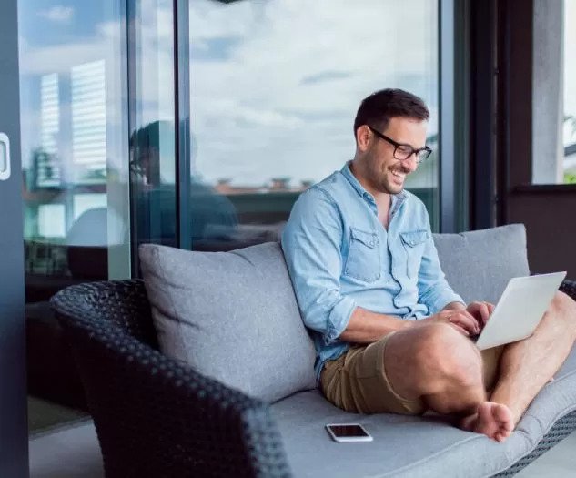 Nasmejan moški v kratkih hlačah in srajci sedi sproščeno na terasi z računalnikom v naročju in dela ali brska po spletu medtem ko je pametni telefon položen ob njem