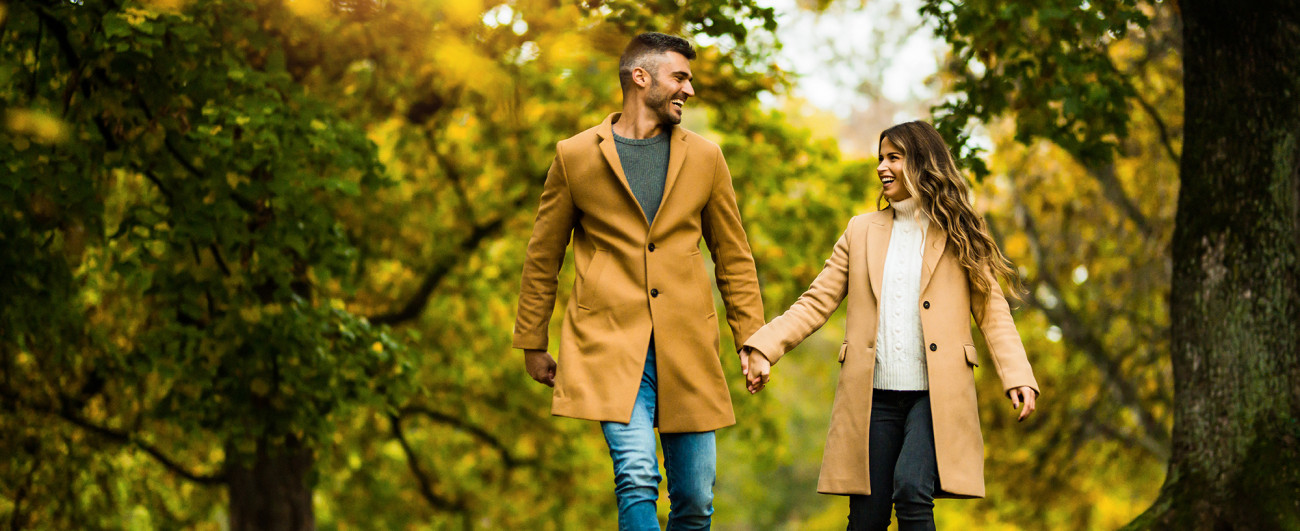 Par se nasmejano drži za roke med sprehodom skozi park obdan z jesenskim zelenjem
