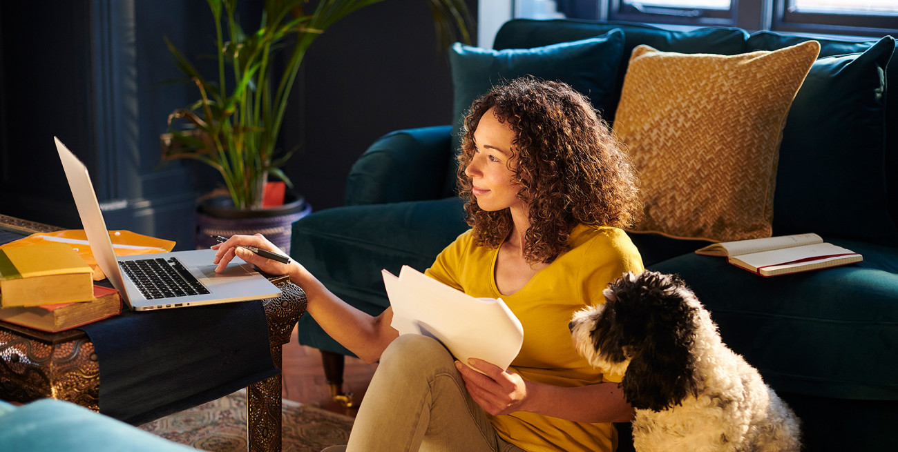 Ženska sedi na tleh v dnevni sobi z dokumenti v roki in dela na prenosniku medtem ko ob njej sedi pes