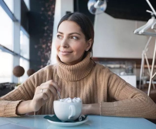 Ženska v toplem puloverju sedi v kavarni ob oknu in z nasmeškom meša vročo čokolado medtem ko zre skozi okno v daljavo