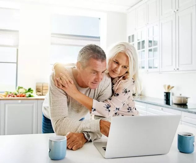 Starejši par stoji objet pred prenosnikom v svetli kuhinji in skupaj gleda v zaslon z zadovoljnim izrazom na obrazu