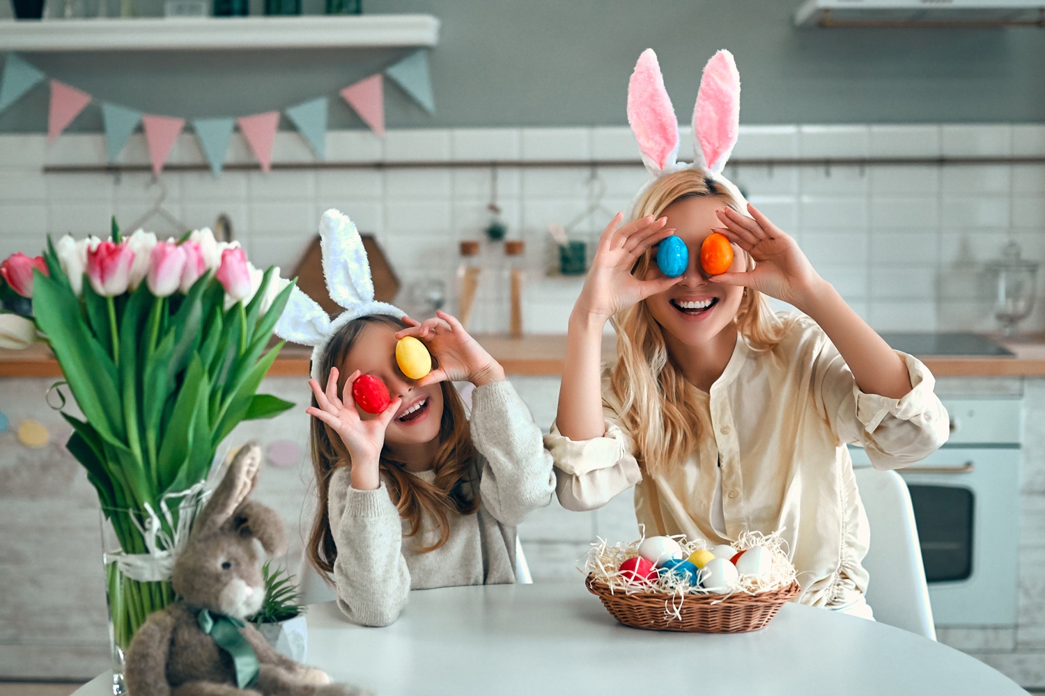 Mama in otrok z zajčjimi ušesi v kuhinji držita barvana velikonočna jajca pred očmi in se smejita